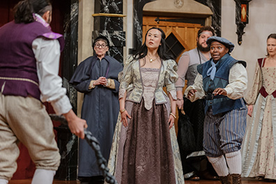 Antipholus of Ephesus with back to camera and holding his rope as across from him the crowd stares at him: Adriana in greentrimed off-green and brown Elizabethan dress, Pinch in Renaissance violet justice robe and glasses calmly standing with hands clenched at his waist, The courtesan in a bodice, dress, and bushy beard, Dromio of Ephesus in blue vest and cap, blue striped knee pants with white stockings, and Luciana in brown and green patterend Elizabethan dress