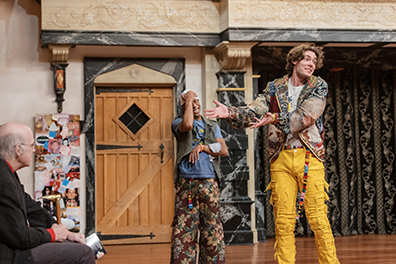 Benvolio has his hand to his white-capped head in embarrassment at Romeo's insult to an audience member sitting on the stage, Romeo's hands pointing in the patron's direction as he looks forward at the rest of theater audience with a teasing expression on his face. 