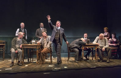 In the center, Hickey in a pinstripe three-piece suit, watch chain across his stomach, high-collar white shirt, strides with right hand upraised preaching as, on either side, the other characters watch, except Hugo asleep at his table.