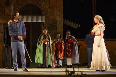 Benedick in pale blue army officers uniform with hands on hilt stands to one side across from Beatrice in yellow layered dress, while the other characters stand in the background in cloaks.